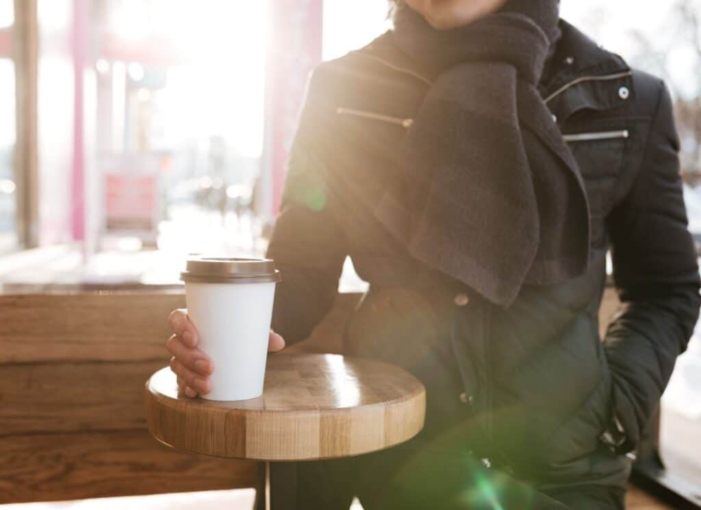 person wearing a jacket holding a cup of coffee that is set on a wooden table outside